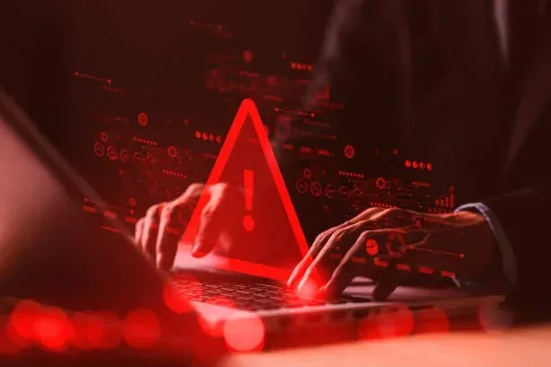 Red warning sign overlay on a person typing on a laptop screen symbolizing cybersecurity threat and hacker warning.
